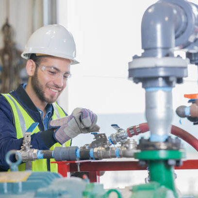 Young adult Hispanic man is working on equipment on pipeline job site for oil and gas company. Engineer is wearing hard hat and observing correct safety procedures.